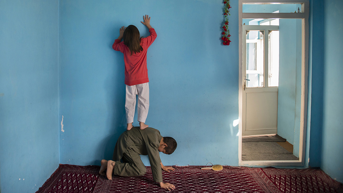 A girl stands on her brother’s back to hang a photo of their father on the wall.